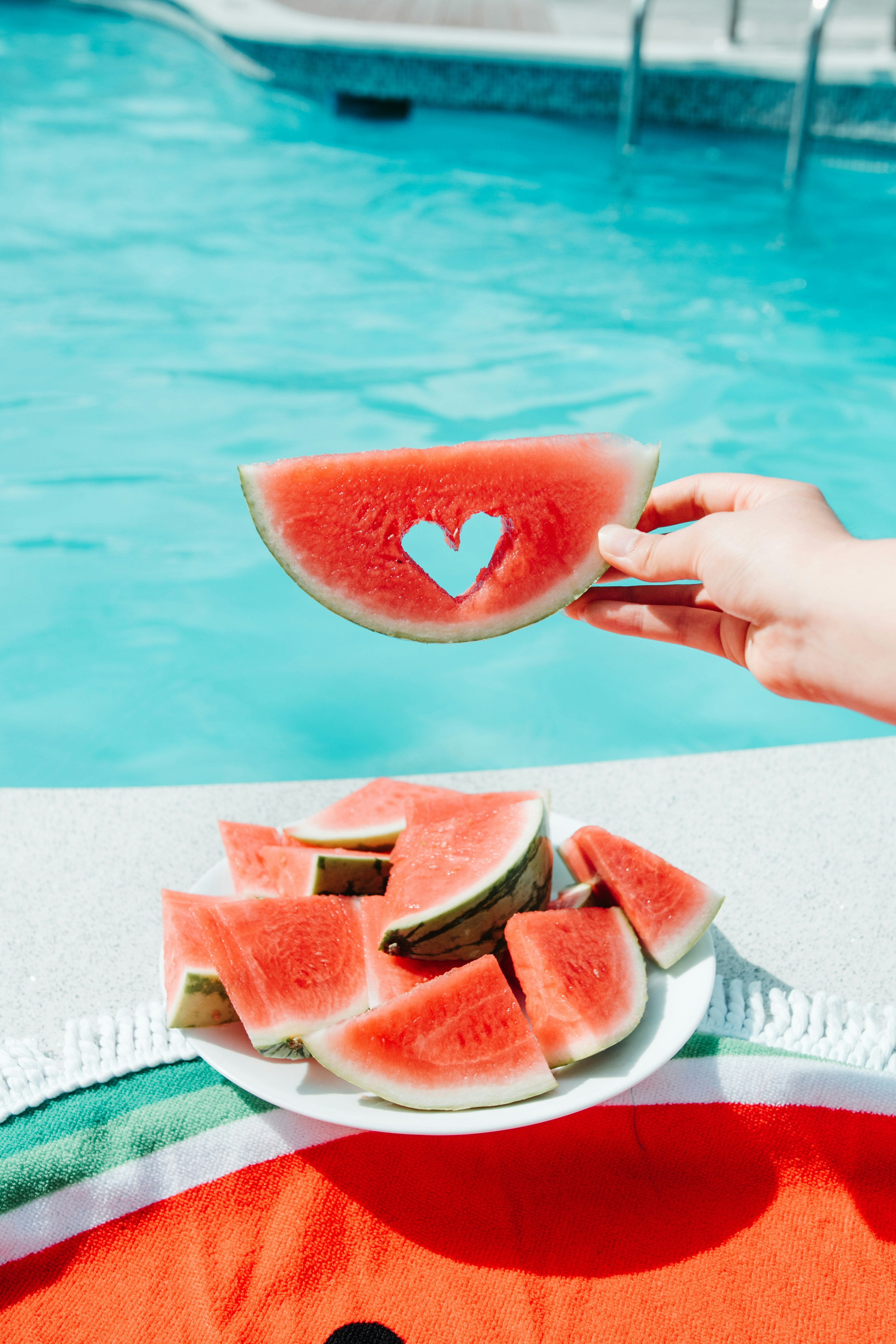 A hand holding a slice of watermelon with a heart-shaped cutout, positioned above a plate of watermelon wedges by a blue swimming pool.